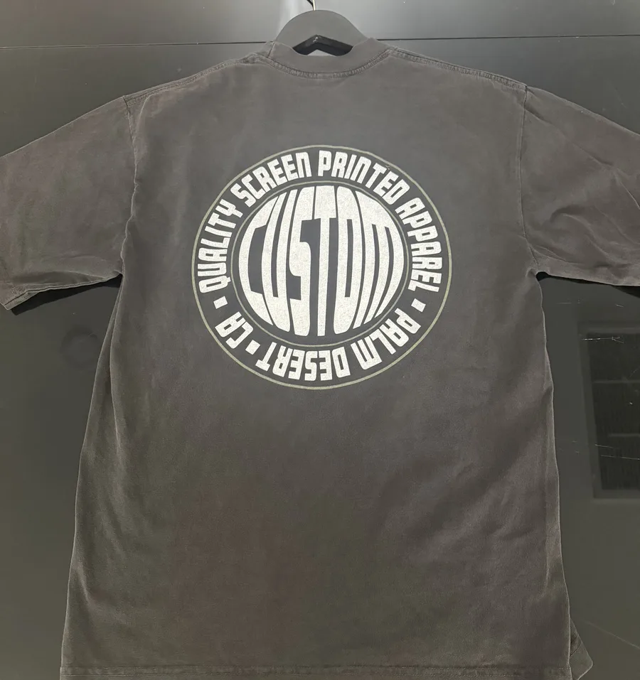A faded black tee laid flat with a large white circular back print reading Custom, Quality Screen Printed Apparel, Palm Desert, CA