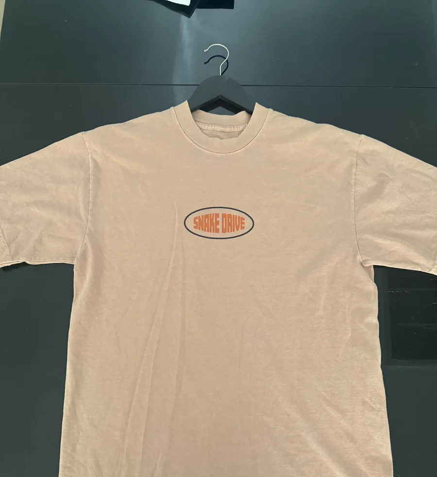 A peach tee laid flat with a small orange and navy oval Snake Drive logo printed on the chest